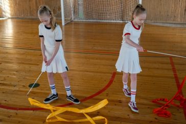 Two girls practice rhythmic gymnastics with ribbons on a wooden indoor court.