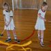 Two girls practice rhythmic gymnastics with ribbons on a wooden indoor court.