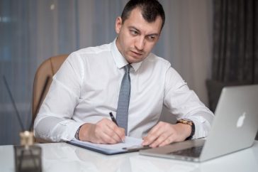 Focused businessman in white shirt writing notes at desk with laptop.